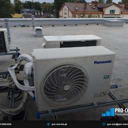 PRO-CON SERWIS Klimatyzacja Wentylacja Rekuperacja - Klimatyzatory Panasonic R32 na dachu budynku, widoczne rury w izolacji, tło stanowią budynki mieszkalne i pochmurne niebo. Montaż na metalowej konstrukcji.