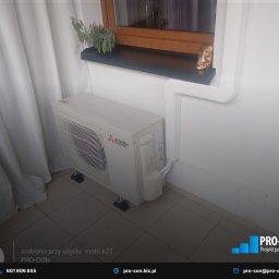 PRO-CON SERWIS Klimatyzacja Wentylacja Rekuperacja - Biały klimatyzator Mitsubishi Electric zamontowany pod oknem z rośliną i dekoracyjną czaszką, widoczne białe rury instalacyjne i zasłona. Minimalistyczny design.