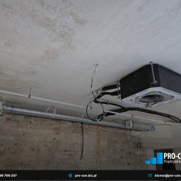 PRO-CON SERWIS Klimatyzacja Wentylacja Rekuperacja - Czarna jednostka klimatyzacyjna zamontowana pod sufitem z widocznymi przewodami i rurami wentylacyjnymi. Surowy mur z cegły w dolnej części kadru.
