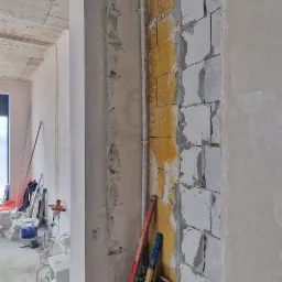 Wnętrze w trakcie remontu, widoczne nieotynkowane ściany z bloków, piony instalacyjne z rur w kolorach czerwonym i niebieskim, poziomice oparte o ścianę, resztki materiałów budowlanych i narzędzia...