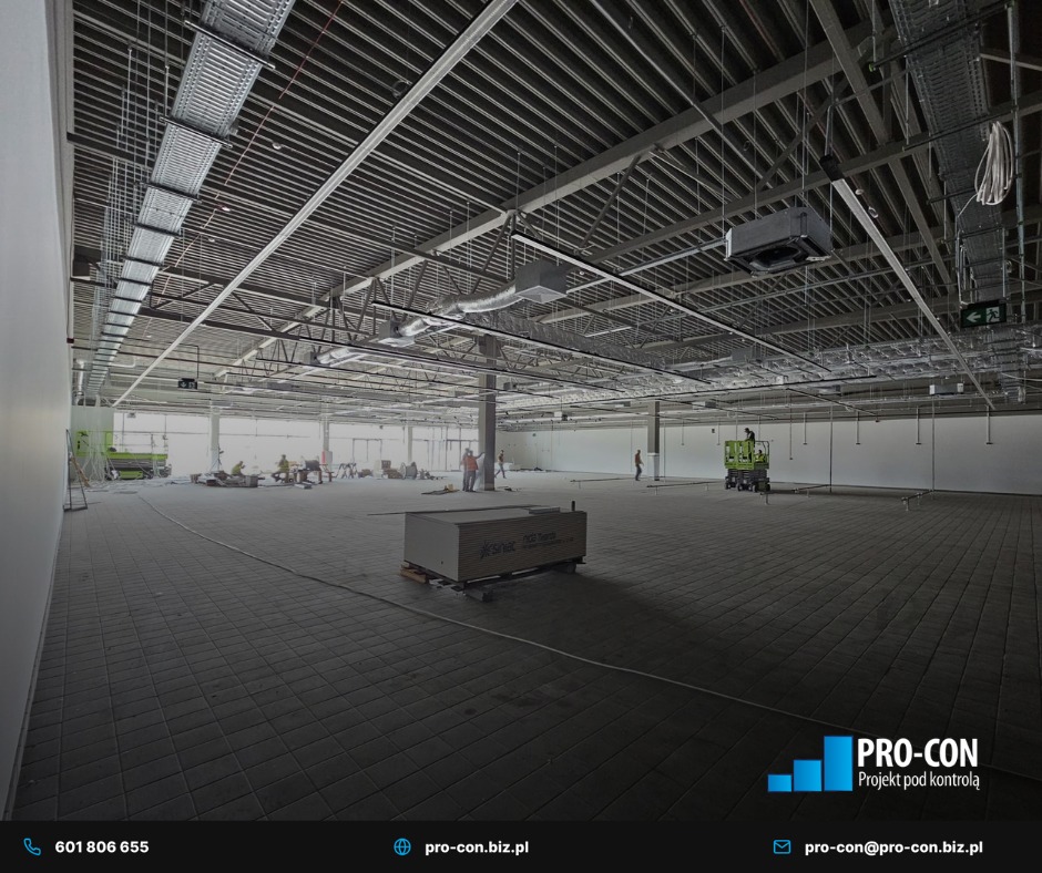 Rozległe wnętrze hali w trakcie instalacji wentylacji pod sufitem. Widoczne kanały wentylacyjne, kable i pracownicy na podnośnikach. Projekt pod kontrolą.