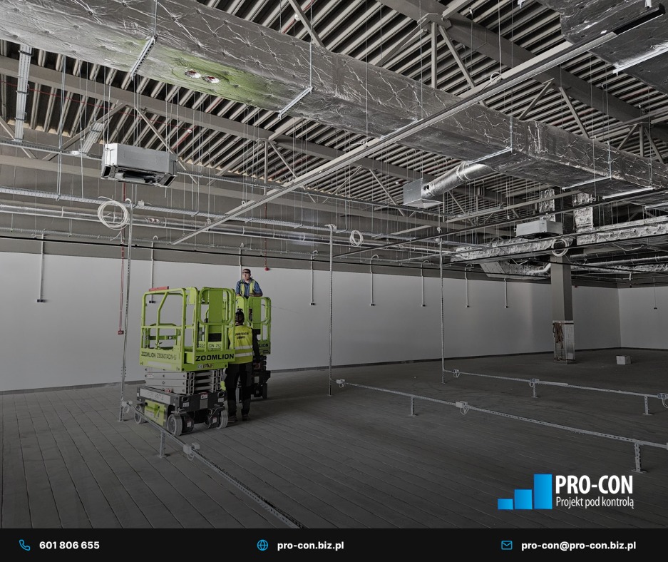 Montaż instalacji wentylacyjnej w hali: pracownicy na podnośnikach montują srebrne kanały pod sufitem. Widoczne elementy konstrukcyjne budynku i oświetlenie.