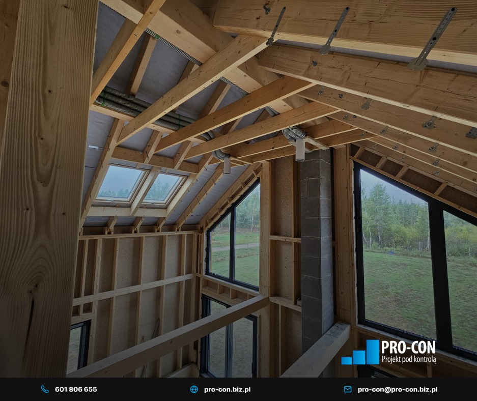 Surowy stan poddasza z widoczną konstrukcją drewnianą, oknami dachowymi i elementami instalacji wentylacyjnej pod stropem. Widok na zewnątrz przez duże okna.