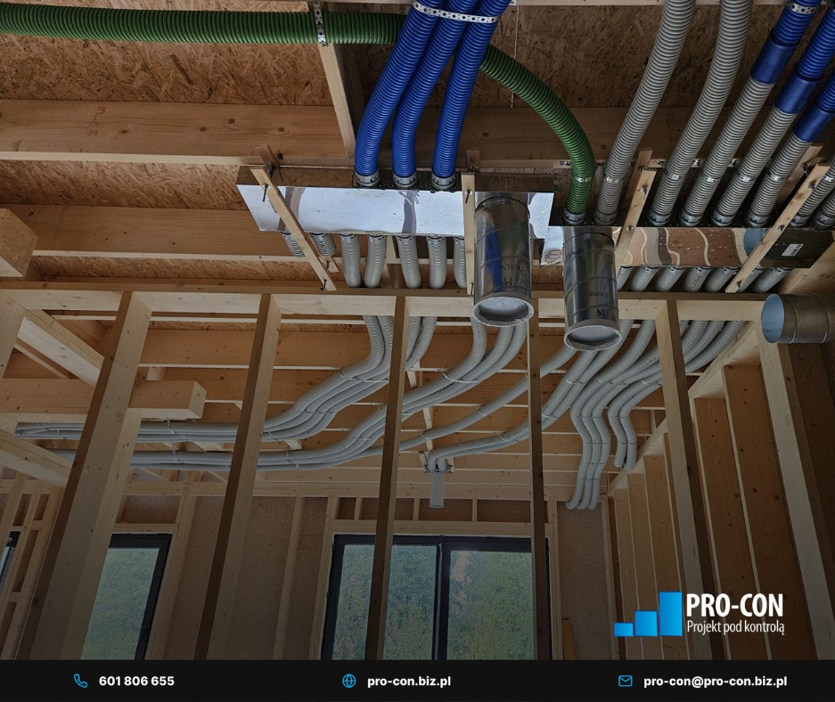 Instalacja wentylacji w surowym budynku: kolorowe rury wentylacyjne (zielone, niebieskie, szare) zamontowane pod drewnianym stropem. Widoczne elementy konstrukcji budynku.