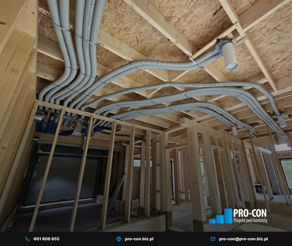 Szare, elastyczne rury wentylacyjne zamontowane pod drewnianym stropem w surowym budynku. Widoczne elementy instalacji i konstrukcja szkieletowa.