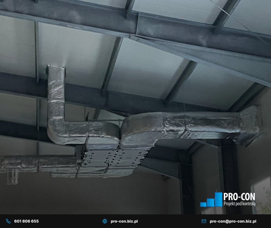 Szare, zaizolowane kanały wentylacyjne pod sufitem hali przemysłowej, widoczne elementy konstrukcji stalowej. Ujęcie z dołu, lekko z prawej strony.