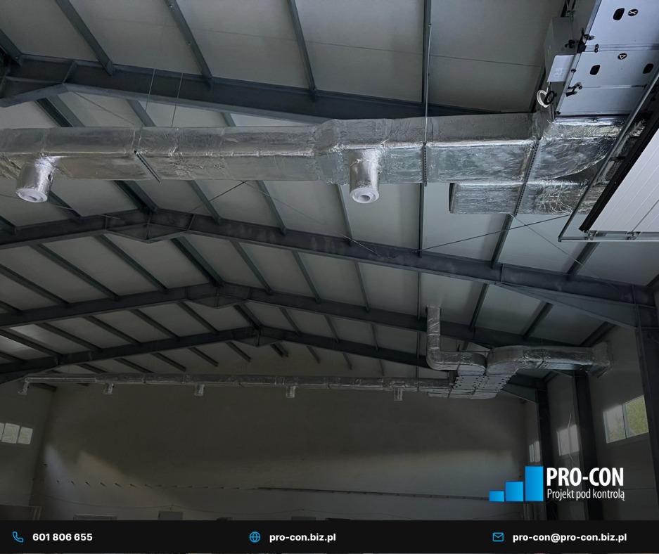 Instalacja wentylacyjna z izolacją w hali przemysłowej, widok z dołu. Metalowa konstrukcja dachu, srebrne rury wentylacyjne z białymi wylotami. Logo firmy w prawym dolnym rogu.