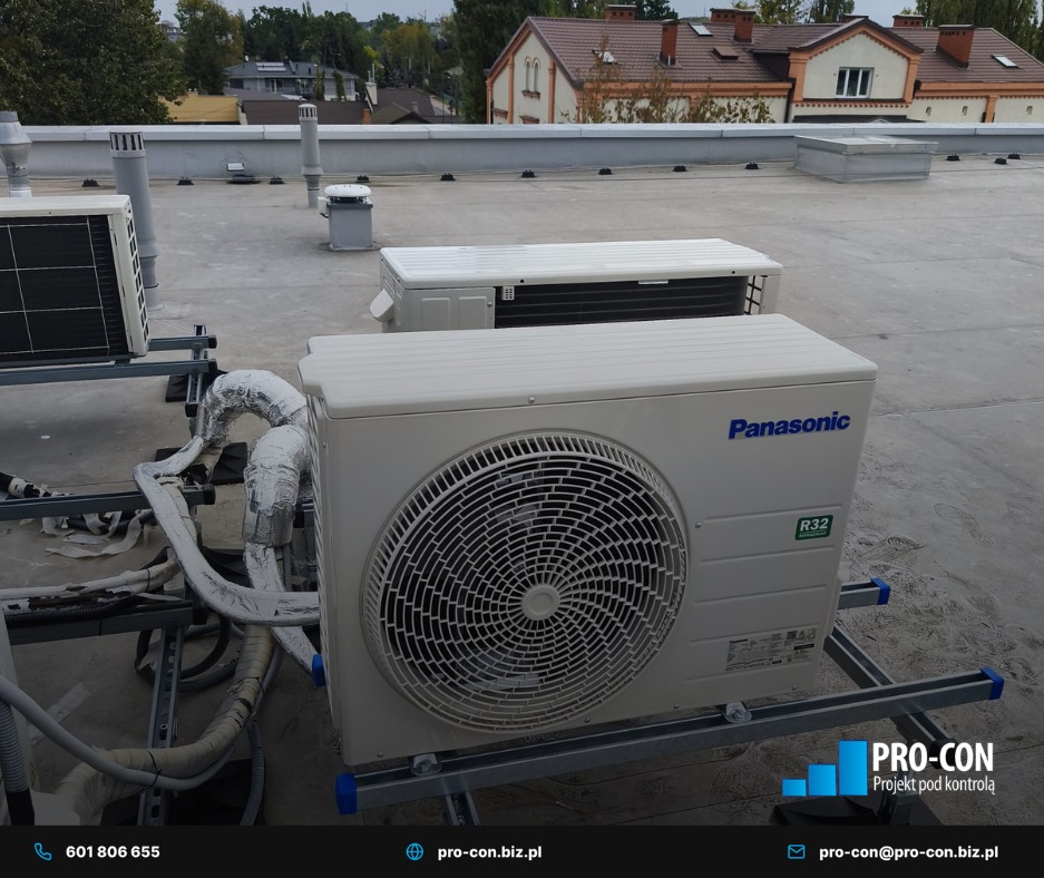 Klimatyzatory Panasonic R32 na dachu budynku, widoczne rury w izolacji, tło stanowią budynki mieszkalne i pochmurne niebo. Montaż na metalowej konstrukcji.