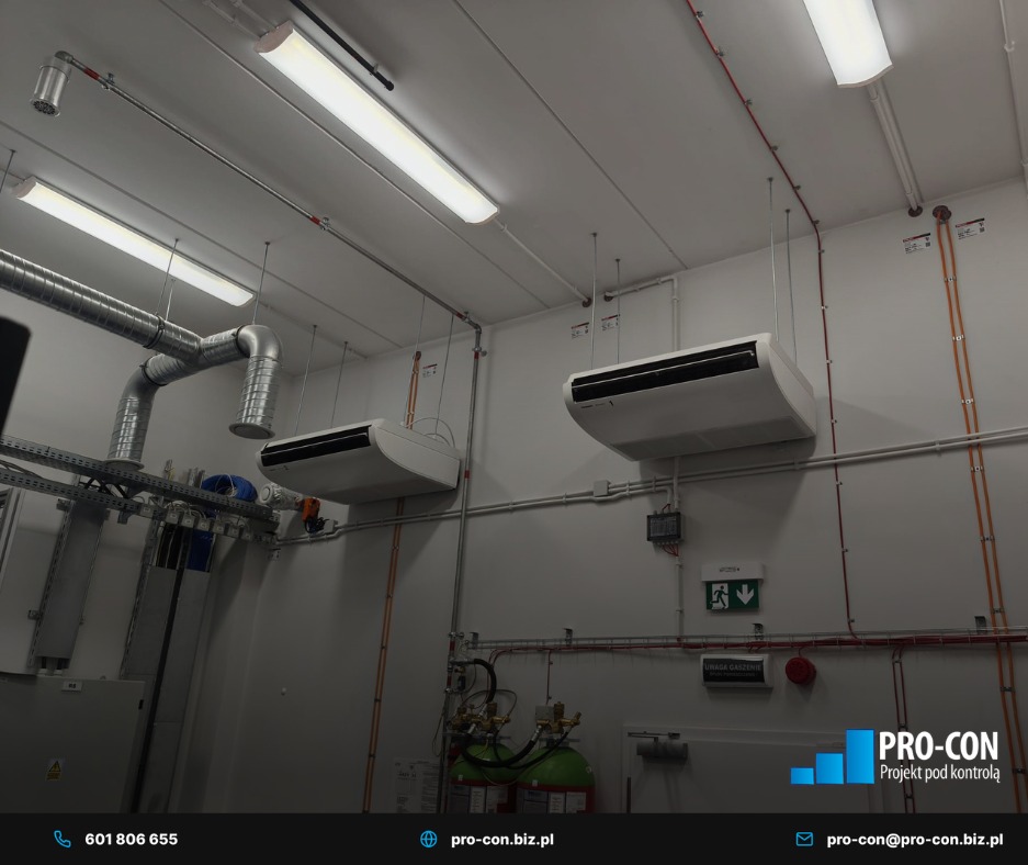 Dwa białe klimakonwektory podwieszone pod sufitem w pomieszczeniu technicznym z widocznymi instalacjami wentylacyjnymi i przeciwpożarowymi.
