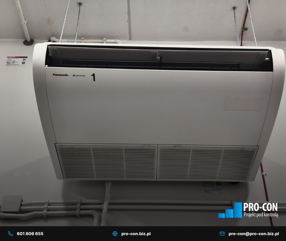 Zamontowana pod sufitem klimatyzacja Panasonic z widocznymi filtrami i numerem '1', instalacja elektryczna w korytku. Szare ściany i sufit. Ujęcie z dołu.