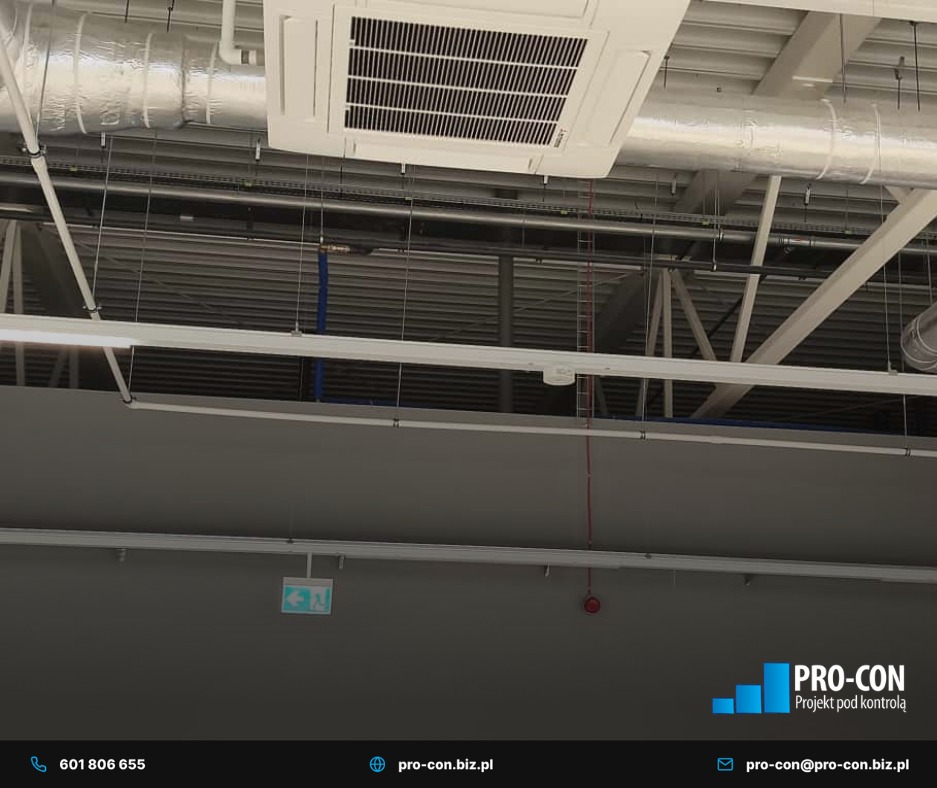 Kasetonowa klimatyzacja sufitowa w hali, widoczne rury wentylacyjne i instalacje. Biała obudowa klimatyzatora z kratką wlotową. Oświetlenie i konstrukcja dachu w tle.