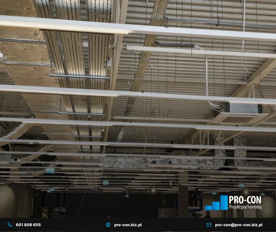 Widok sufitu hali z zamontowaną instalacją wentylacyjną i klimatyzacyjną. Widoczne kanały wentylacyjne, rury i klimatyzator. Oświetlenie liniowe.