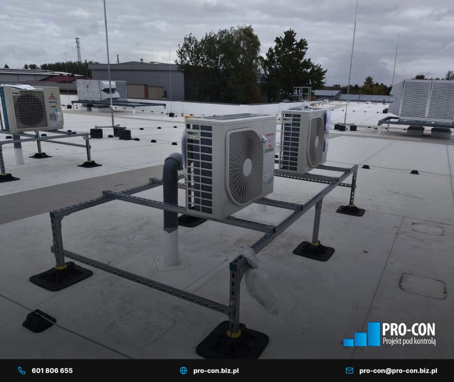 Klimatyzatory na dachu budynku, zamontowane na metalowej konstrukcji wsporczej. Widoczne elementy instalacji i tło z zielenią. Ujęcie z niskiego kąta.