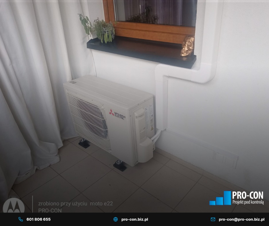 Biały klimatyzator Mitsubishi Electric zamontowany pod oknem z rośliną i dekoracyjną czaszką, widoczne białe rury instalacyjne i zasłona. Minimalistyczny design.