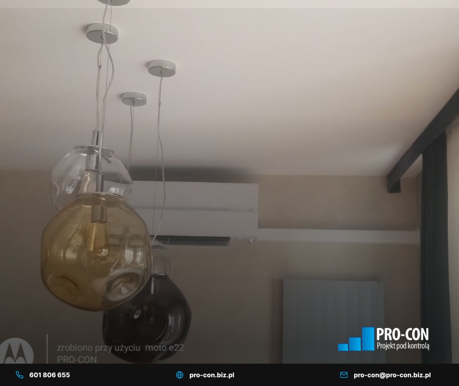 Biała klimatyzacja ścienna w jasnym wnętrzu, widoczna spod dekoracyjnych lamp wiszących. Minimalistyczny design, neutralna kolorystyka ścian.