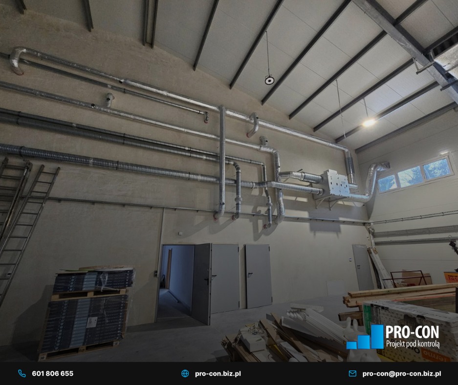 Wnętrze hali z widoczną instalacją wentylacyjną pod sufitem, rury prowadzone wzdłuż ściany, drabina oparta o ścianę, paleta z materiałami na podłodze.