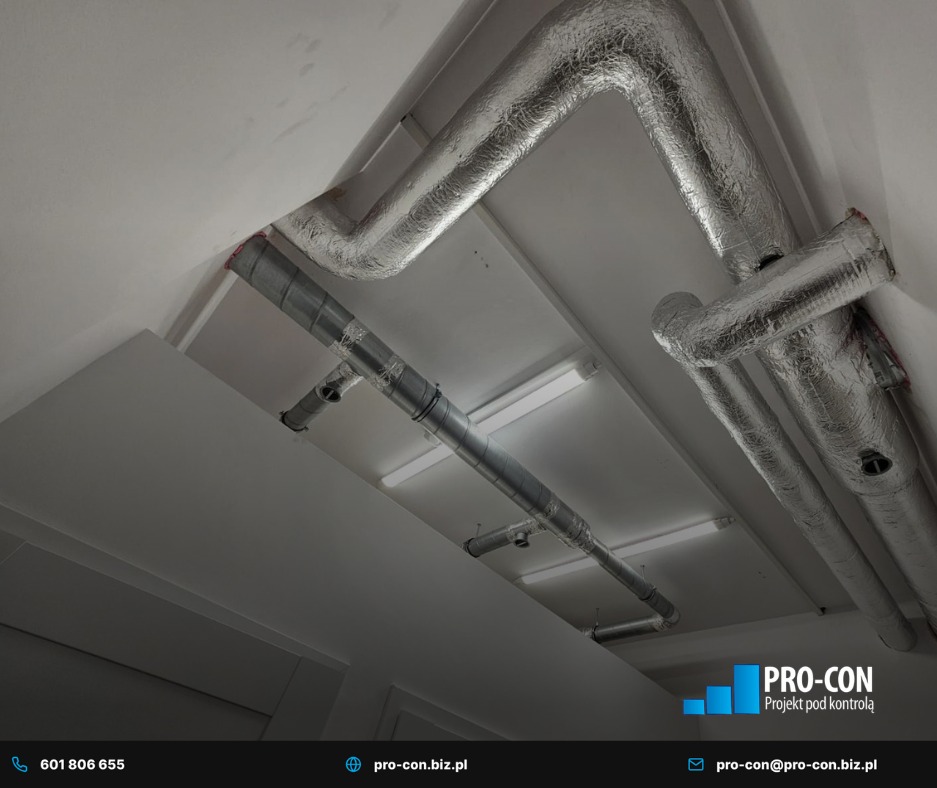 Instalacja wentylacyjna pod sufitem w Grodzisku Mazowieckim. Widoczne rury z izolacją termiczną i elementy mocujące. Oświetlenie sufitowe w tle.