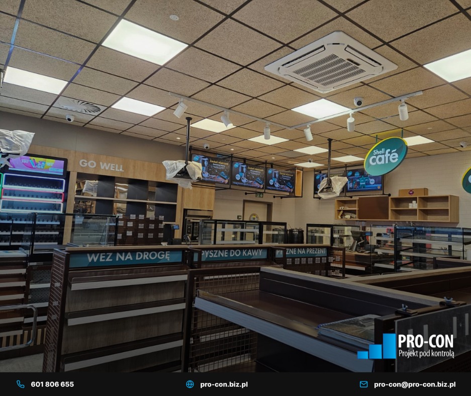 Kasetonowa klimatyzacja sufitowa w lokalu handlowym, widoczna instalacja oświetleniowa i kamery monitoringu. Widok na wnętrze sklepu z regałami i logo Shell Cafe.