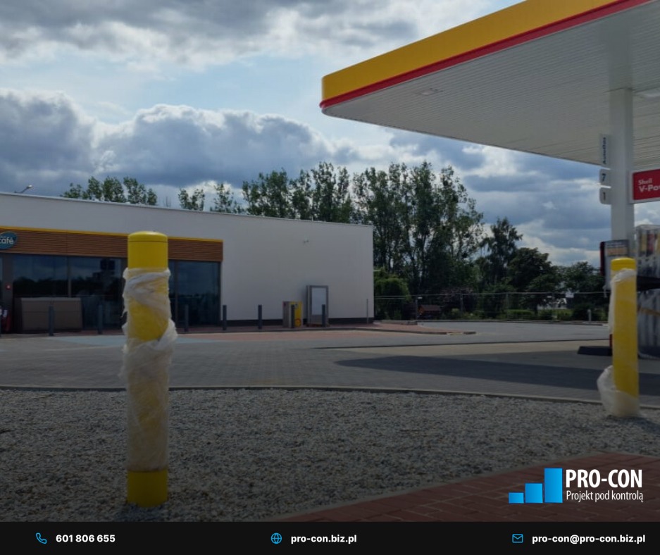 Stacja paliw Shell z żółtymi słupkami ochronnymi, częściowo owiniętymi folią. Widok na budynek stacji i zadaszenie dystrybutorów w pochmurny dzień.