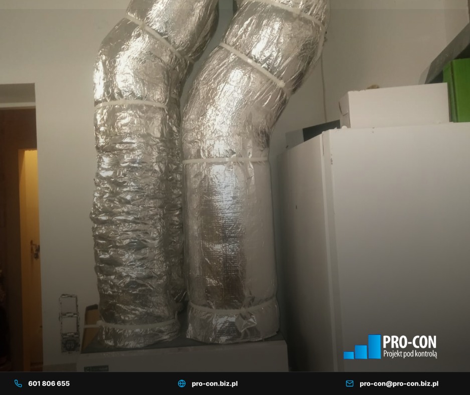 Dwie izolowane, aluminiowe rury wentylacyjne, przymocowane białymi opaskami, w pomieszczeniu z białymi ścianami i urządzeniem. Widoczne logo firmy w prawym dolnym rogu.