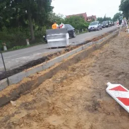 Przygotowanie podłoża pod krawężniki na poboczu drogi; widoczny pracownik w pomarańczowej kamizelce i samochody w tle.