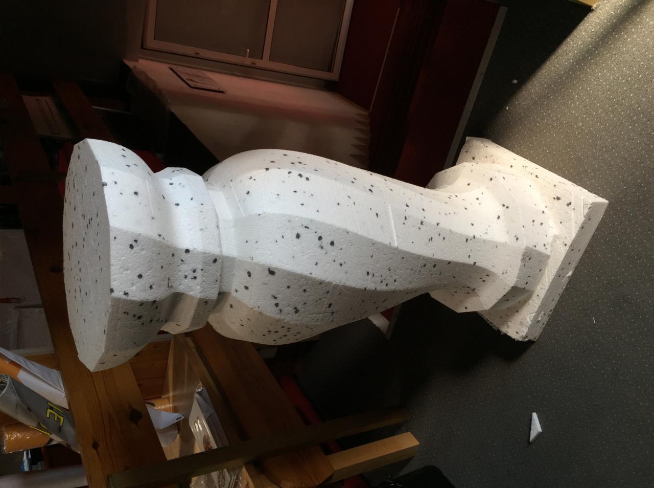 Styropianowy element dekoracyjny przypominający kolumnę, leżący na boku, z widoczną strukturą materiału i nieregularnymi czarnymi plamkami.