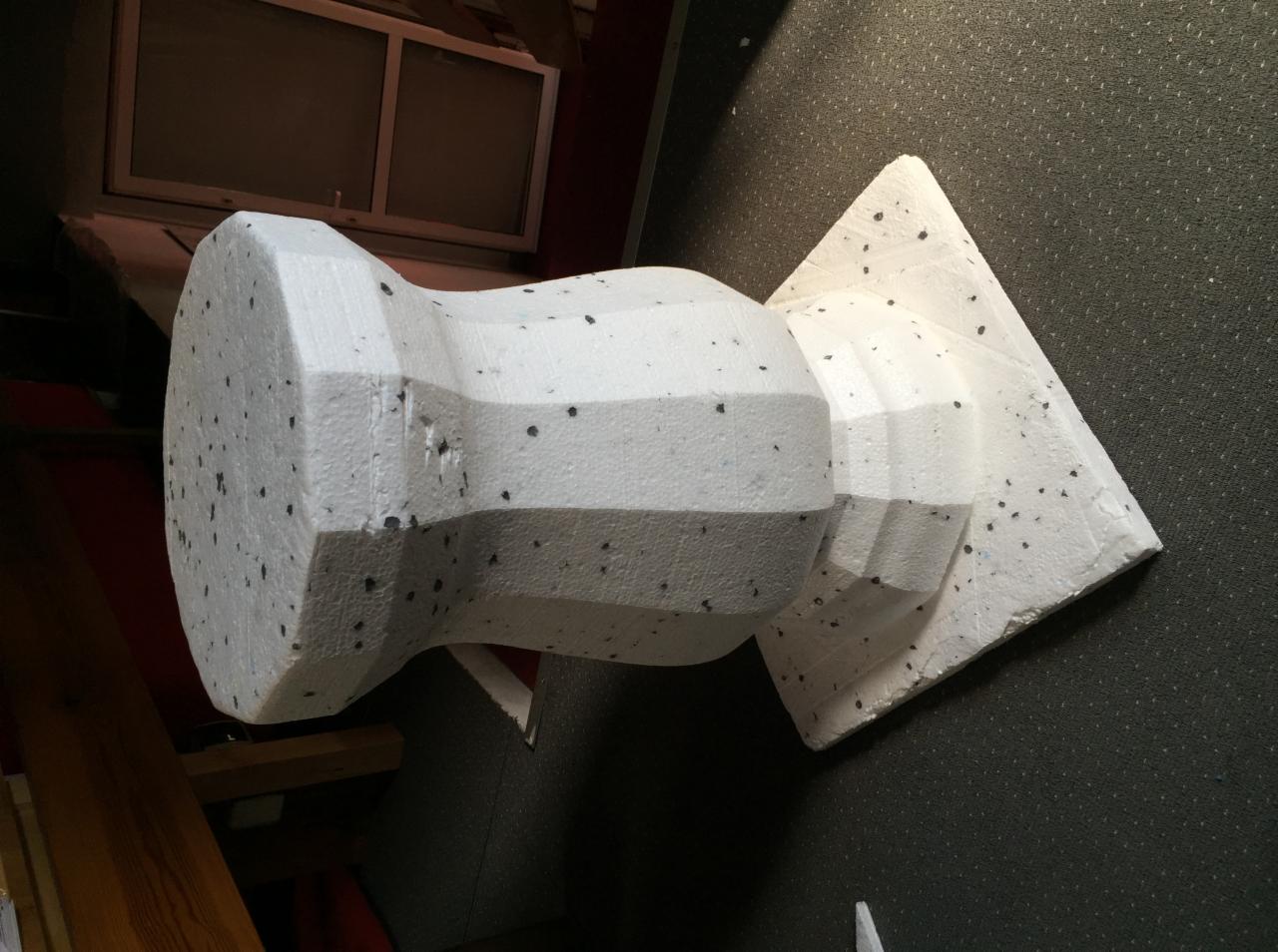 Niedokończona rzeźba z białego styropianu o nieregularnych kształtach, leżąca na szarym podłożu, z widocznymi fragmentami wnętrza pomieszczenia w tle.