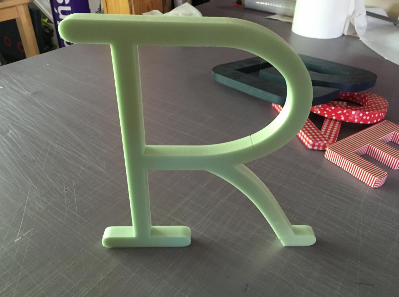 Jasnozielona, trójwymiarowa litera R z tworzywa sztucznego na metalowym blacie, w tle inne litery w różnych wzorach. Usługi reklamowe w Legionowie.