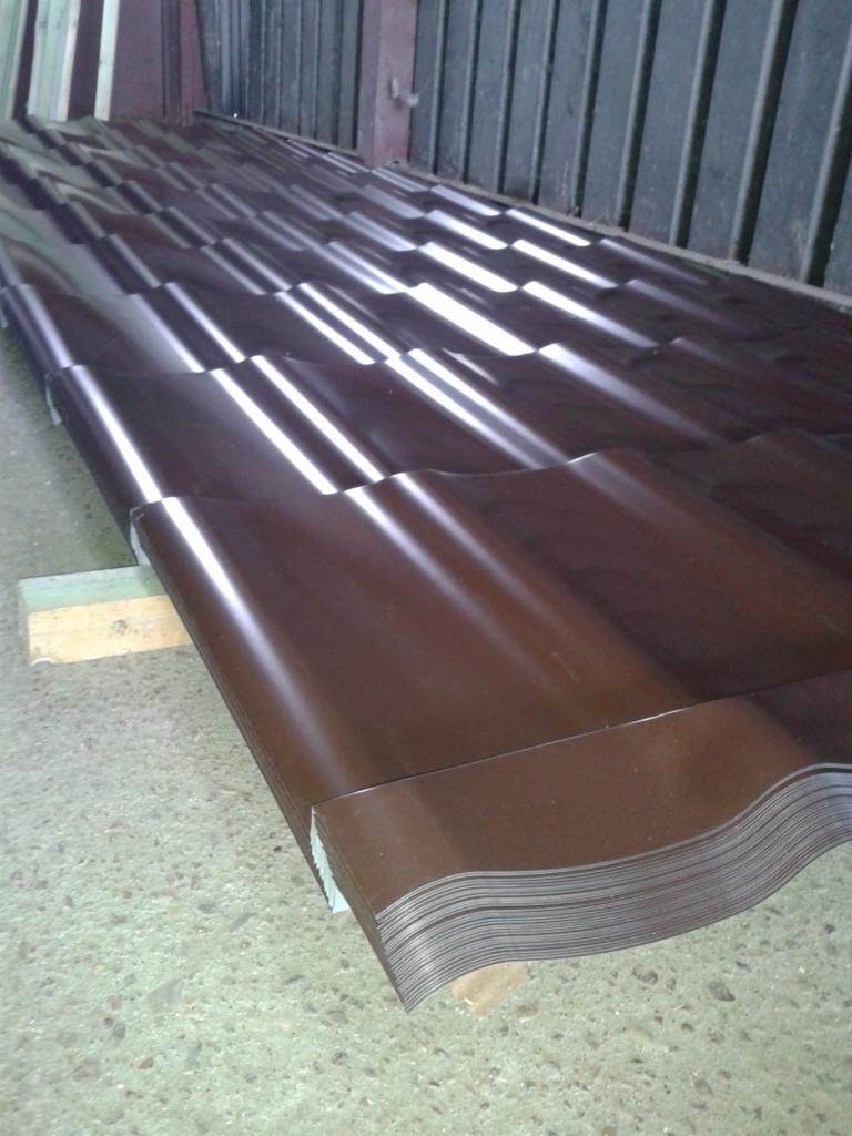 Stos brązowej, profilowanej blachy na drewnianych podkładkach, widoczne odbicia światła na powierzchni, tło stanowią metalowe panele.