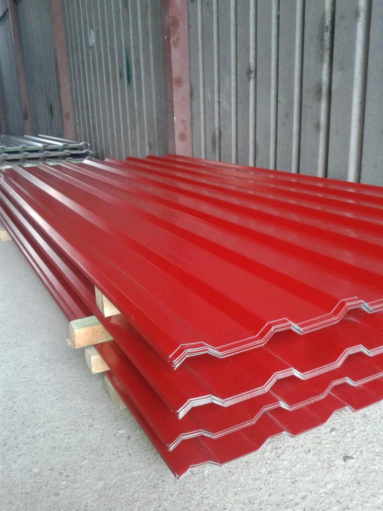 Stos czerwonych arkuszy profilowanych na drewnianych legarach, widoczny profil sinusoidalny, tło stanowi blaszana ściana magazynu.