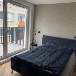 Budekspert - Sypialnia w neutralnych barwach z łóżkiem z ciemną pościelą i widokiem na balkon przez duże okno. Na ścianie kinkiety. Minimalistyczny wystrój.