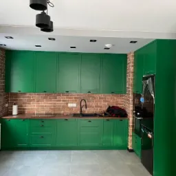 Nowoczesna kuchnia z zielonymi szafkami, ceglaną ścianą i czarnym sprzętem AGD. Widoczne okno i szara podłoga. Unikalne oświetlenie sufitowe.