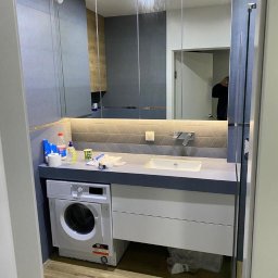 Budekspert - Nowoczesna łazienka z pralką pod umywalką i lustrami szafkowymi. Szara kolorystyka, drewniana podłoga. Widoczne akcesoria i odbicie osoby w lustrze.
