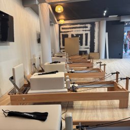 Budekspert - Jasne wnętrze studia pilates z rzędem drewnianych reformerów. W tle widoczna ściana w trakcie wykończenia i lampy wiszące. Minimalistyczny design.