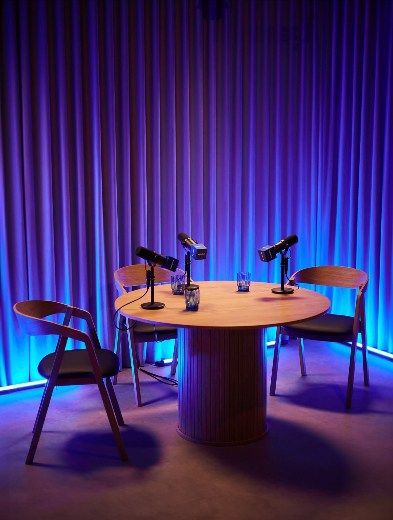 Studio podcastowe: okrągły stół z mikrofonami i szklankami, otoczony krzesłami na tle niebieskich zasłon i neonowego oświetlenia.