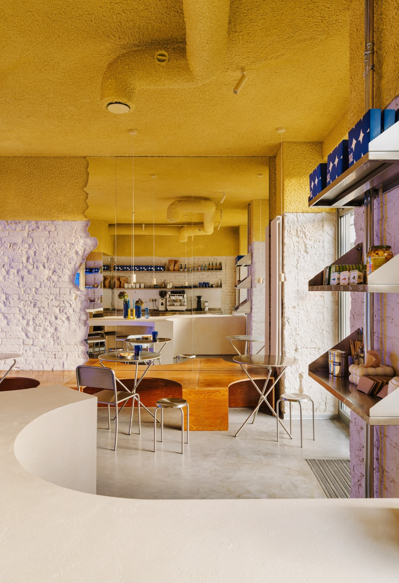 Wnętrze kawiarni z żółtym, teksturowanym sufitem, białymi ścianami z cegły i drewnianymi elementami. Metalowe stoliki i krzesła podkreślają nowoczesny design.