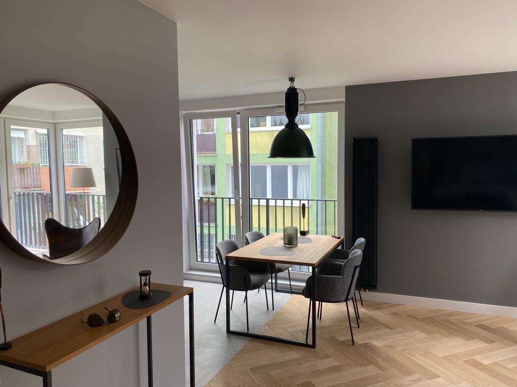 Nowoczesne wnętrze jadalni z parkietem w jodełkę, okrągłym lustrem i minimalistycznym stołem z szarymi krzesłami. Widok na balkon i budynek zza okna.
