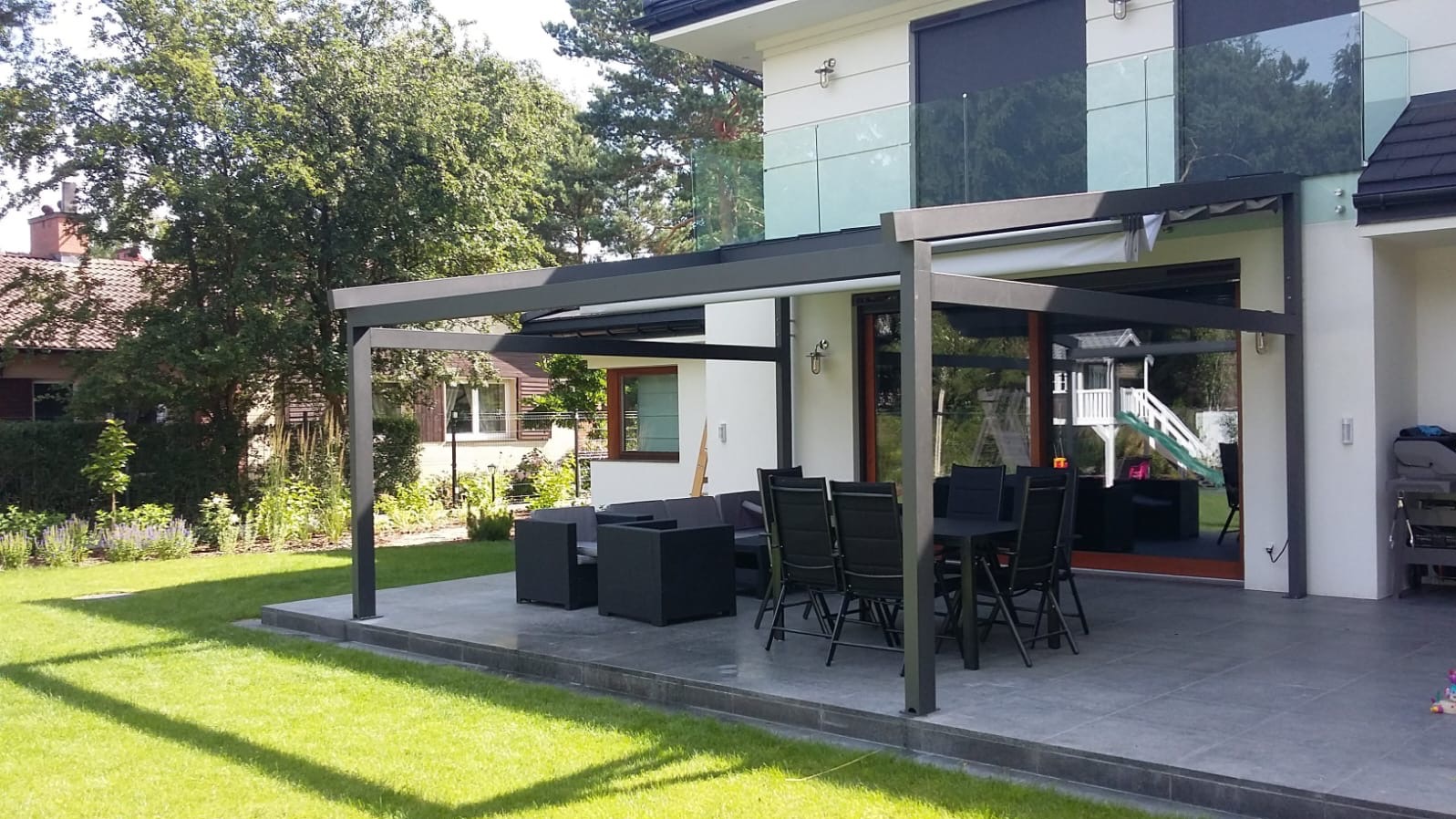 Nowoczesna pergola z szarymi meblami ogrodowymi na tarasie z płytek. W tle zielony trawnik i budynek z oknami. Widoczne refleksy w szklanej balustradzie.