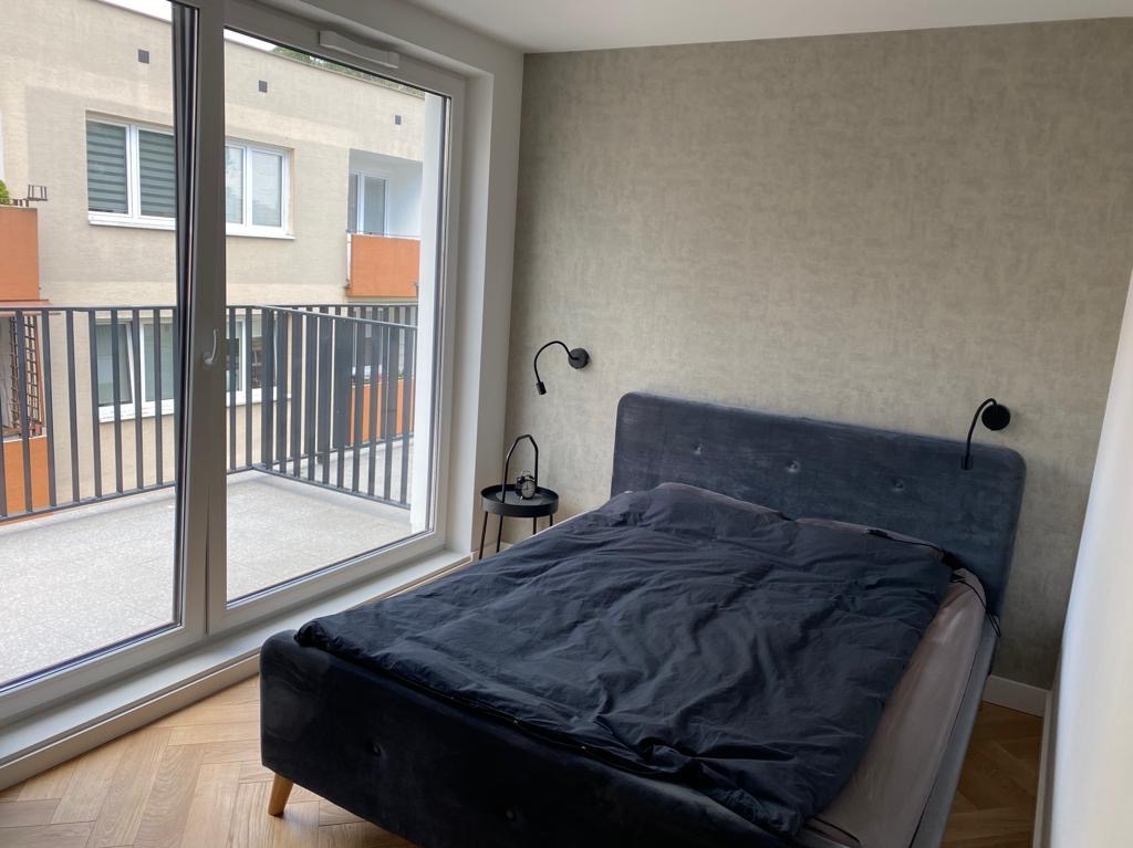 Sypialnia w neutralnych barwach z łóżkiem z ciemną pościelą i widokiem na balkon przez duże okno. Na ścianie kinkiety. Minimalistyczny wystrój.