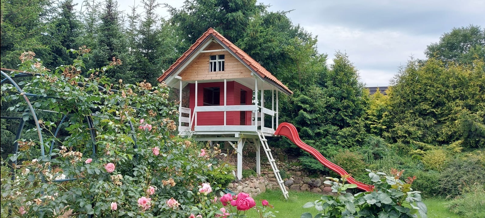 Domek na drzewie z czerwoną zjeżdżalnią, otoczony kwitnącymi różami i zielenią. Drewniana konstrukcja na palach, widoczna drabina i dach z dachówką.