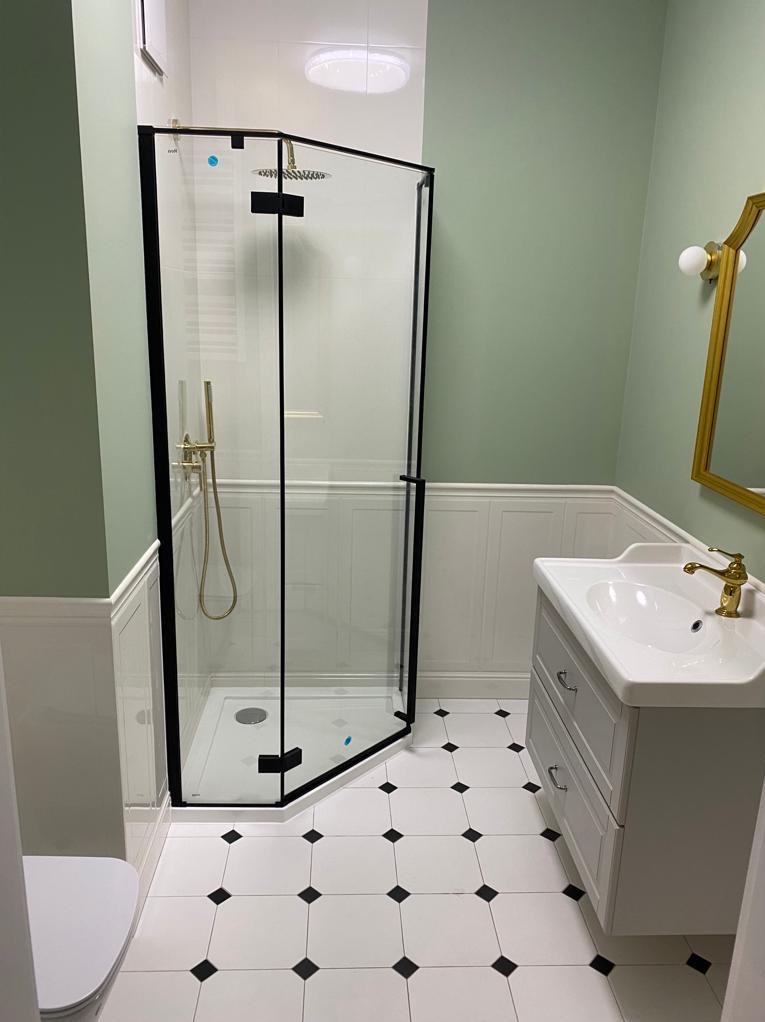 Wykończona łazienka w stylu retro: kabina prysznicowa z czarnymi ramami, biała umywalka z armaturą w kolorze złota i biało-czarna podłoga w karo.