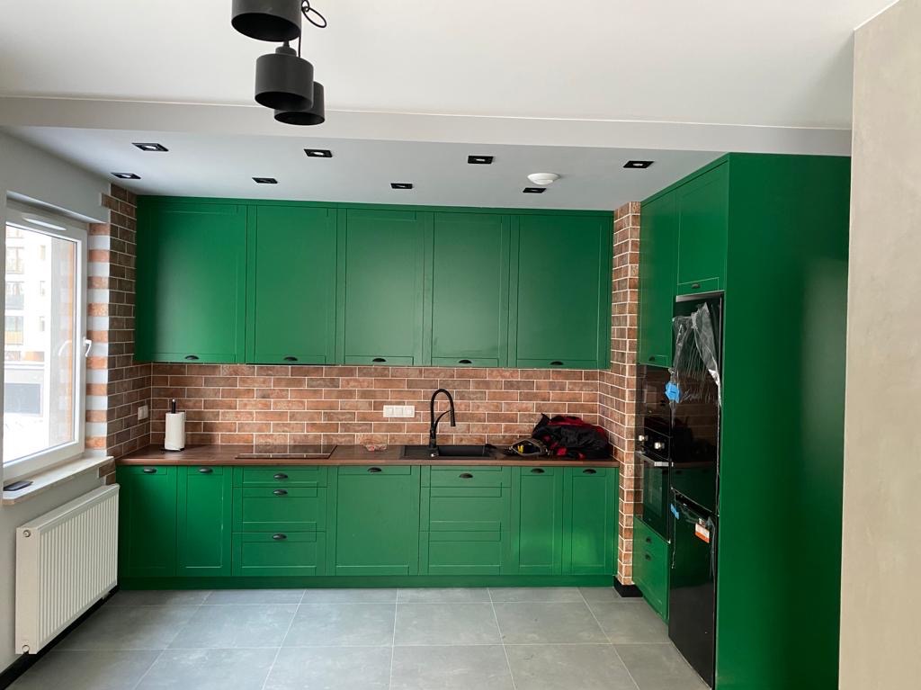 Nowoczesna kuchnia z zielonymi szafkami, ceglaną ścianą i czarnym sprzętem AGD. Widoczne okno i szara podłoga. Unikalne oświetlenie sufitowe.