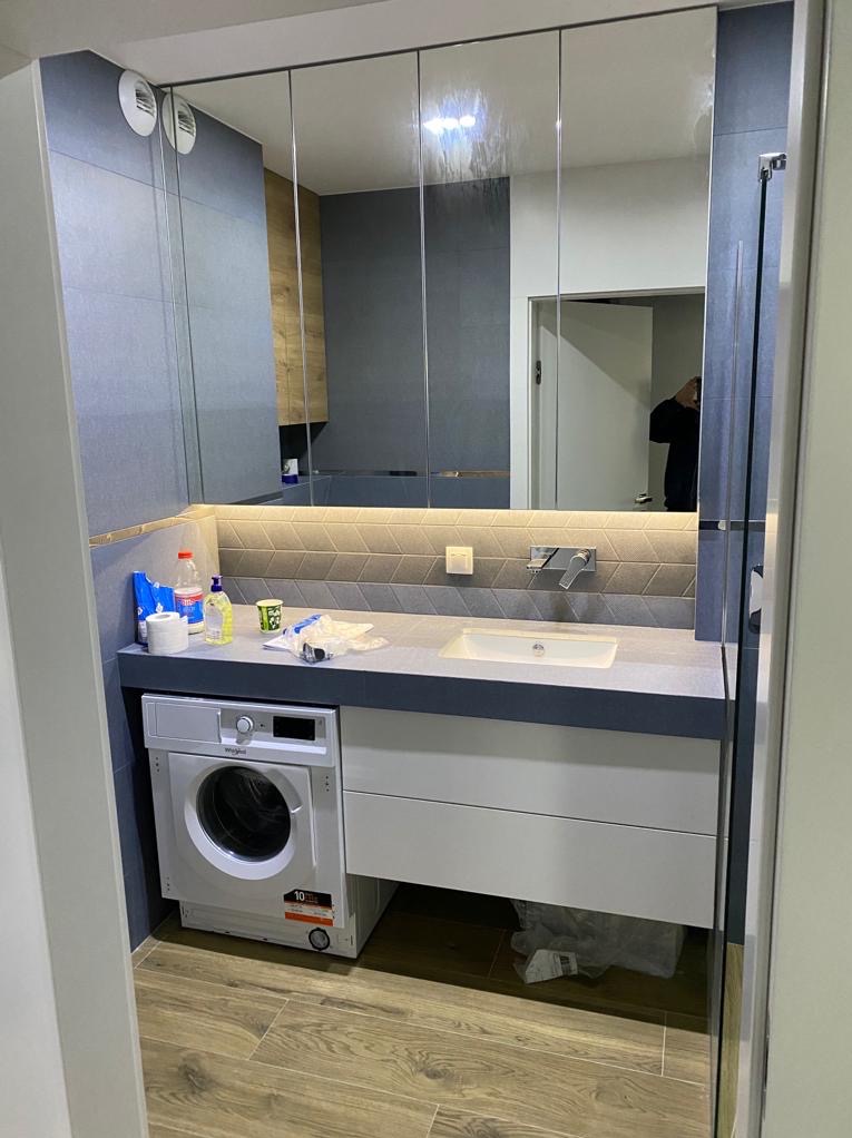 Nowoczesna łazienka z pralką pod umywalką i lustrami szafkowymi. Szara kolorystyka, drewniana podłoga. Widoczne akcesoria i odbicie osoby w lustrze.