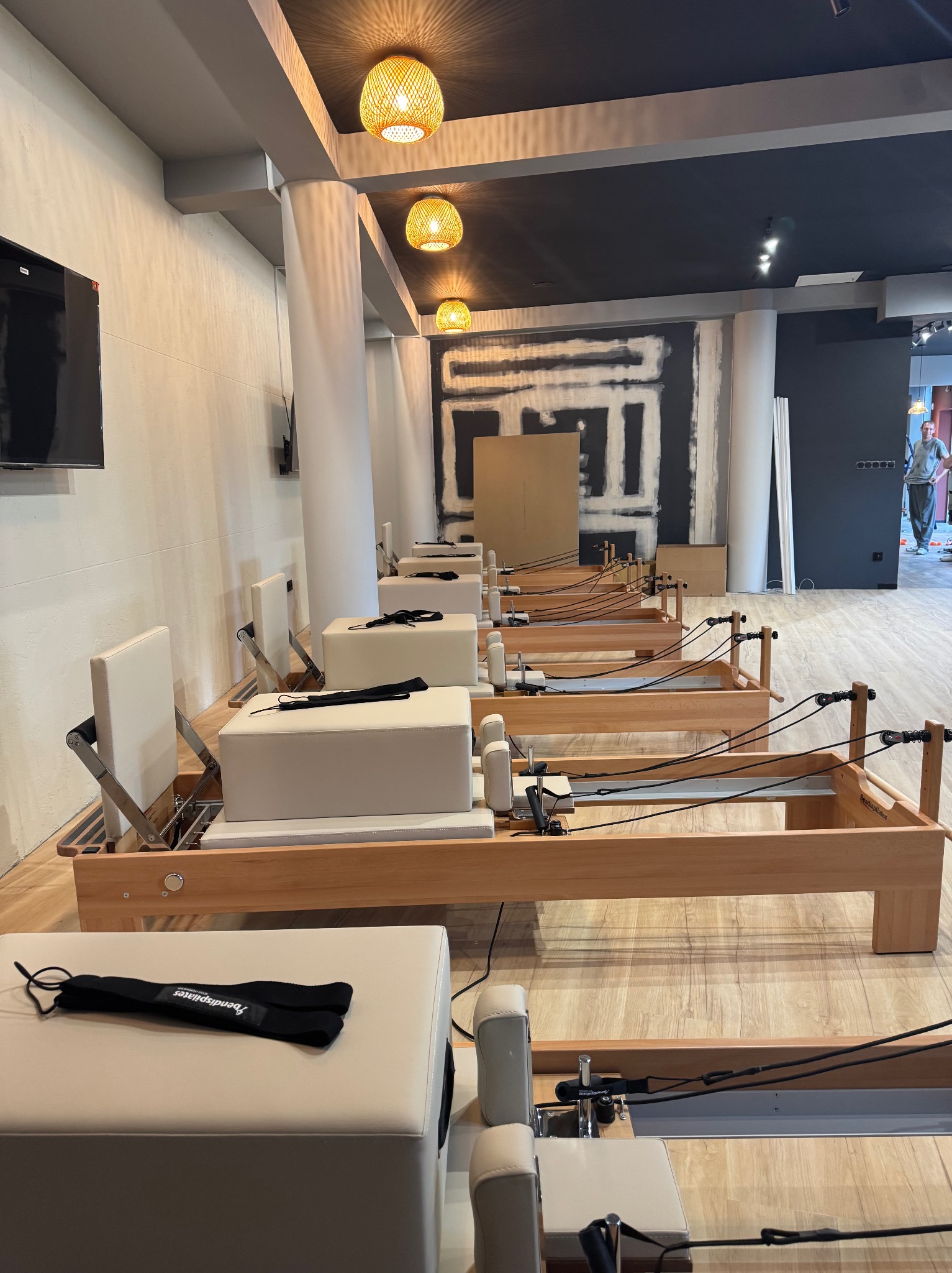 Jasne wnętrze studia pilates z rzędem drewnianych reformerów. W tle widoczna ściana w trakcie wykończenia i lampy wiszące. Minimalistyczny design.