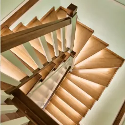 Drewniane schody zabiegowe z podświetleniem LED, widok z góry. Brązowe poręcze z białymi słupkami na tle jasnych ścian.