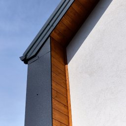 SHINIK GLOBAL Sp. z o. o. - Fragment elewacji domu z drewnianym wykończeniem i metalową obróbką blacharską na tle błękitnego nieba w słoneczny dzień.
