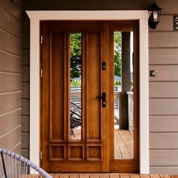 SHINIK GLOBAL Sp. z o. o. - Drewniane drzwi wejściowe z przeszkleniami, osadzone w białej ramie, na tle szarej elewacji z drewnianą podłogą. Widoczny fragment krzesła.