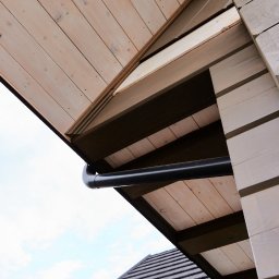 SHINIK GLOBAL Sp. z o. o. - Fragment elewacji drewnianej z rynną w kolorze czarnym, widok z dołu na tle nieba. Widoczny fragment dachu z ciemną dachówką.