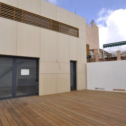 SHINIK GLOBAL Sp. z o. o. - Nowoczesny balkon z drewnianą podłogą i jasną elewacją z poziomymi żaluzjami. Widoczne drzwi balkonowe i fragment sąsiedniego budynku.