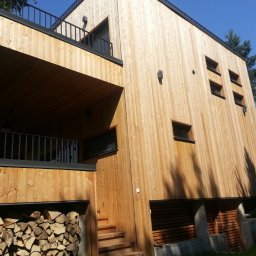 SHINIK GLOBAL Sp. z o. o. - Nowoczesny dom z elewacją z jasnego drewna, widok z dołu. Elementy metalowe w kolorze czarnym. Składowane drewno pod balkonem. Minimalistyczny styl.